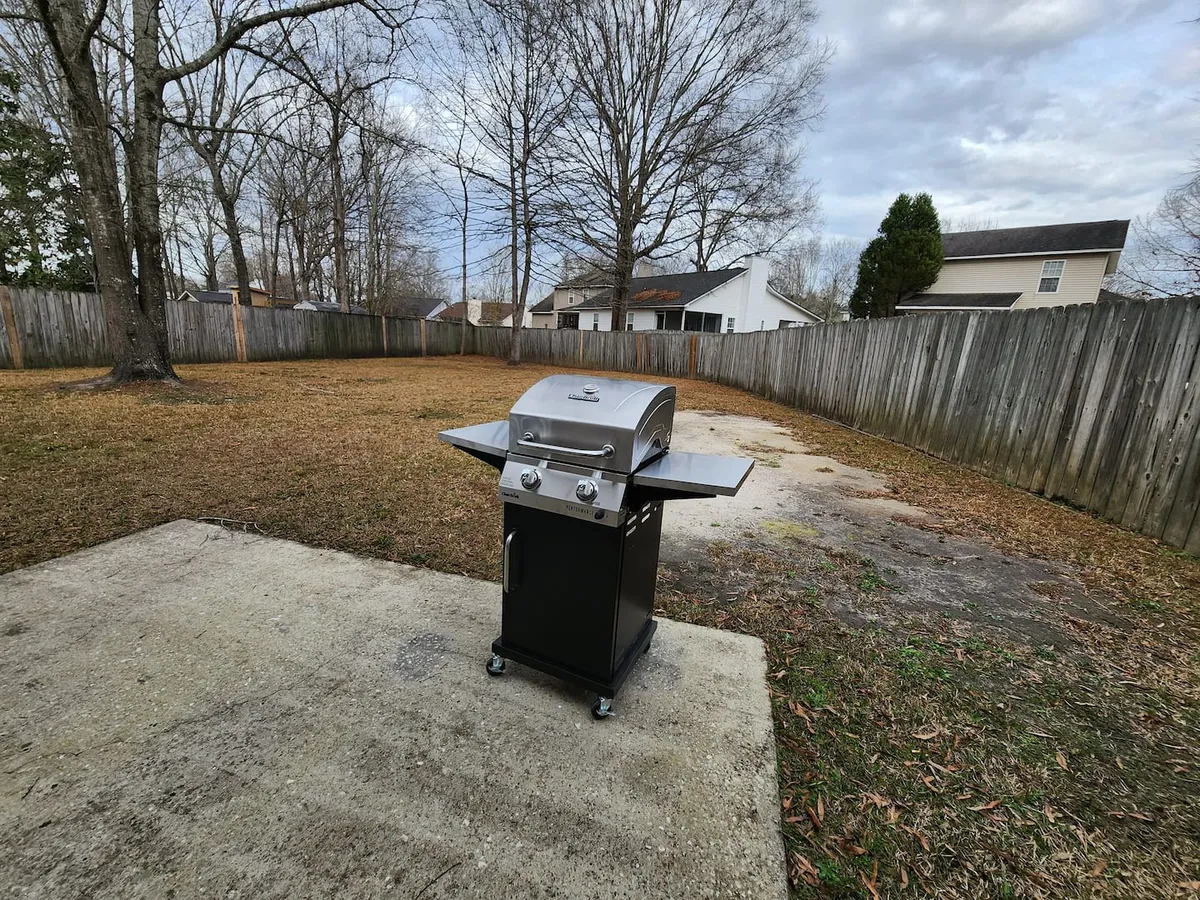 A fenced backyard area with a grill, open green space, and seating options, ideal for casual outdoor dining and socializing. The space feels private and ready for a laid-back barbecue or evening relaxation.
