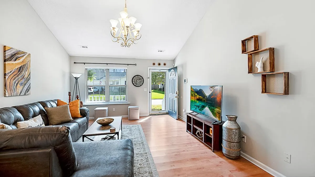 A cozy and inviting living room featuring a comfortable sofa, a coffee table, a large television, and decorative shelving near the front door. The space is bright and uncluttered, with a layout that encourages relaxation or social gatherings.