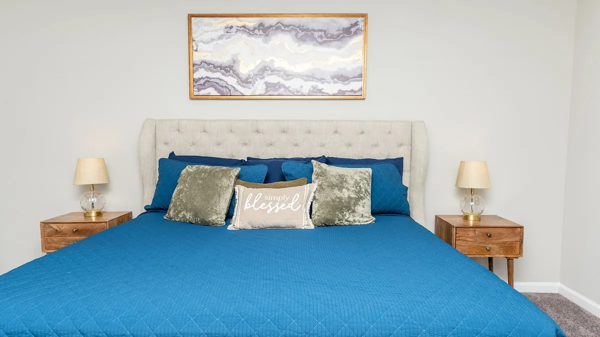 A tranquil bedroom featuring a bed with a blue quilt, decorative pillow with text, matching nightstands with lamps, and abstract artwork above the headboard. The room is softly lit, evoking a restful ambiance.