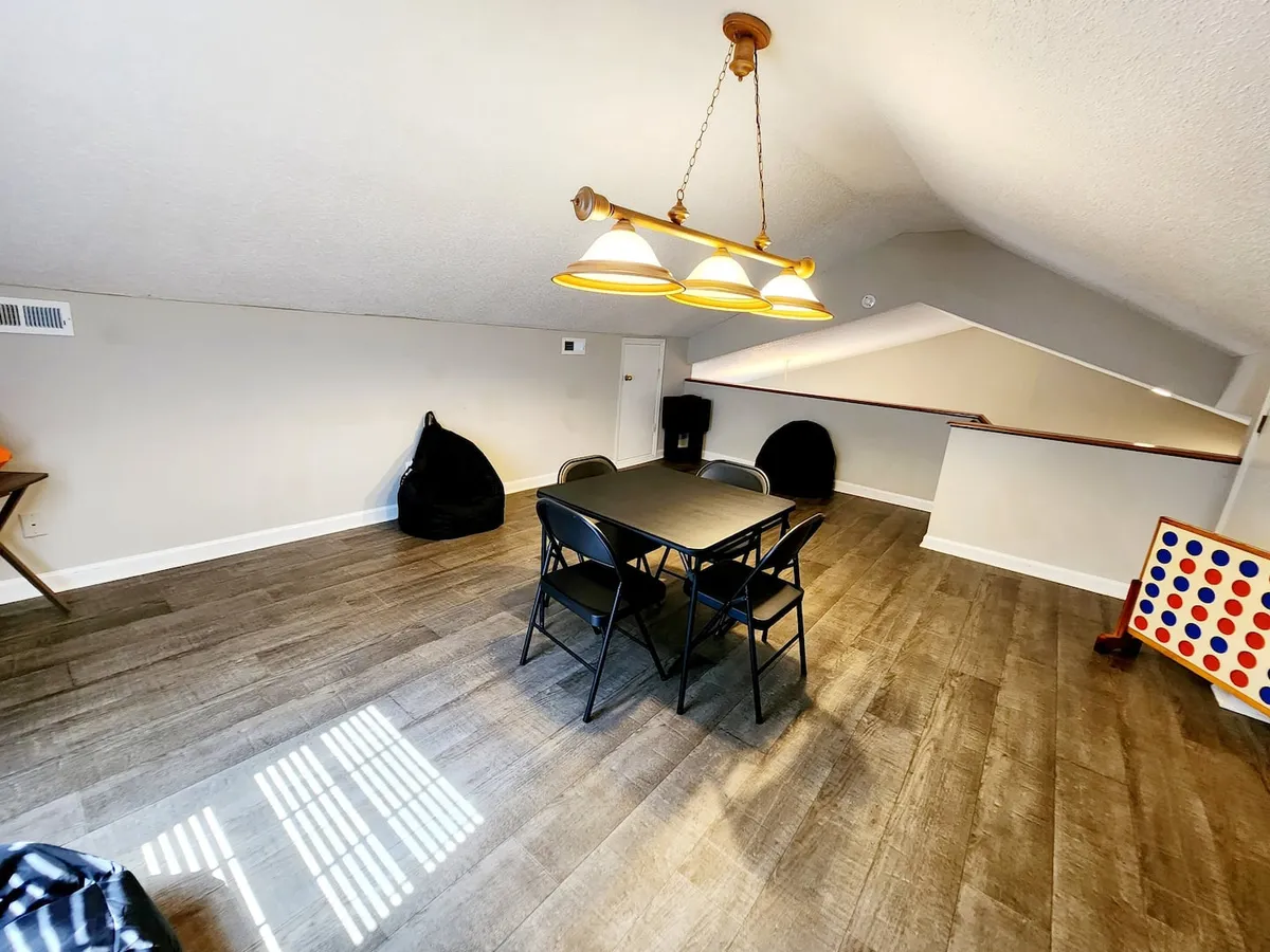 A bright, inviting dining and game space featuring a dining table with comfortable seating, vibrant bean bags, a playful Connect Four game, and a modern light fixture. The setting is casual and designed for group interaction, with elements that encourage both dining and friendly competition.