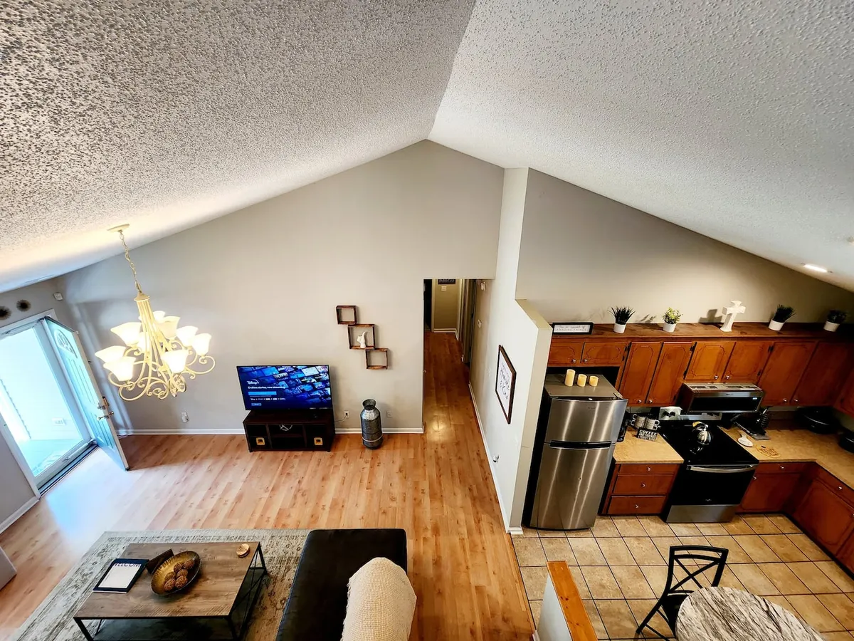 An open-plan living space with a plush sofa, a modern chandelier, a TV perfectly positioned for movie nights, and a view of the adjacent kitchen area. The room is well-lit and designed for family gatherings or entertaining guests.