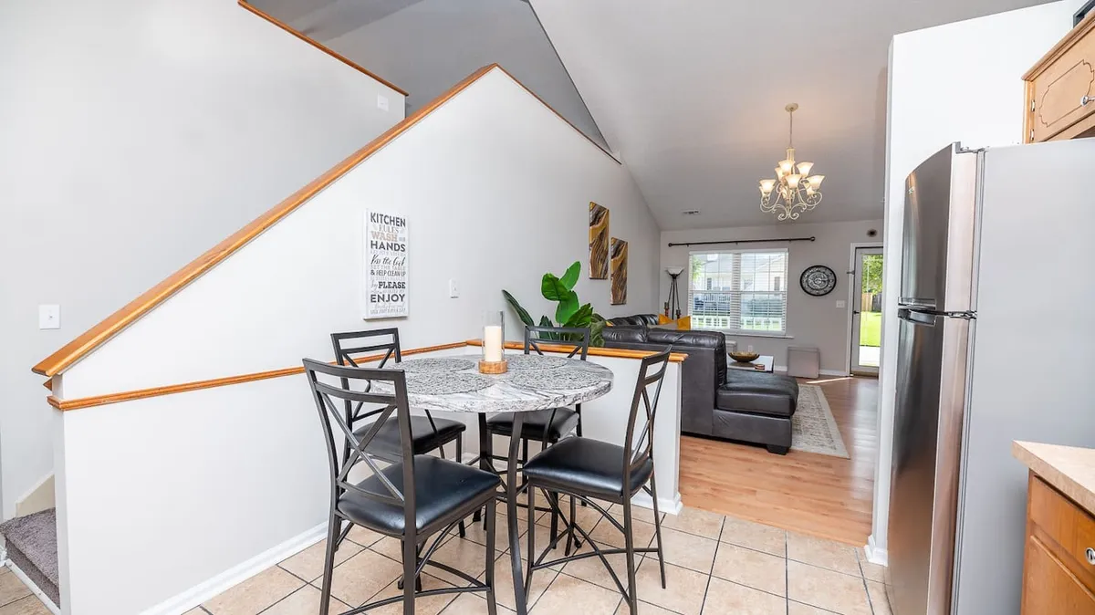A bright, welcoming interior dining and living area includes a round dining table, comfortable couches, a leafy green plant, and a large window letting in natural light. The open layout is perfect for group meals, casual meetings, and relaxation.