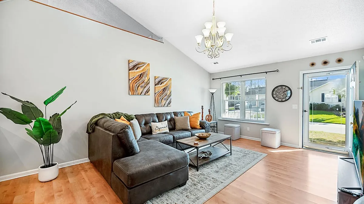 The image showcases a cozy living room with a plush sofa, stylish coffee table, tasteful art pieces on the walls, and a large window that fills the space with natural light. The setting invites guests to relax, socialize, or enjoy a peaceful moment with a book by the window.